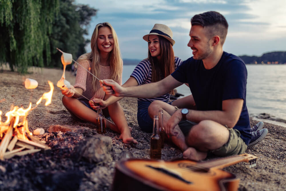 young friends camping on beach gather around fire and roast marshmellows
